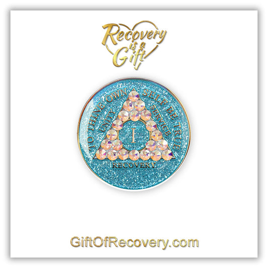 1 year deep sea aqua glitter AA medallion adorned with 21 genuine Aurora Borealis crystals representing the different colors and stages of our new lives, to thine own self be true, unity, service, recovery are embossed with 14k gold-plated brass and sealed with resin for a glossy finish that is scratch proof and will last, seen on a white 3x3 card with recovery is a gift going through an open heart at the top and giftofrecovery.com on the bottom, both in the color gold.