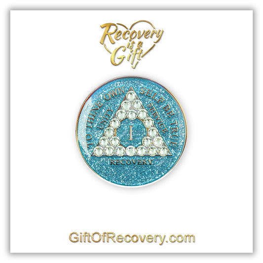 1 year deep sea Aqua glitter AA medallion adorned with 21 diamond CZ genuine crystals in the shape of the triangle, to thine own self be true, unity, service, recovery are embossed with 14k gold-plated brass and sealed with resin for a lasting finish, featured on a white 3x3 card, recovery is a gift at the top going through an open heart and giftofrecovery.com at the bottom, both in the color gold.