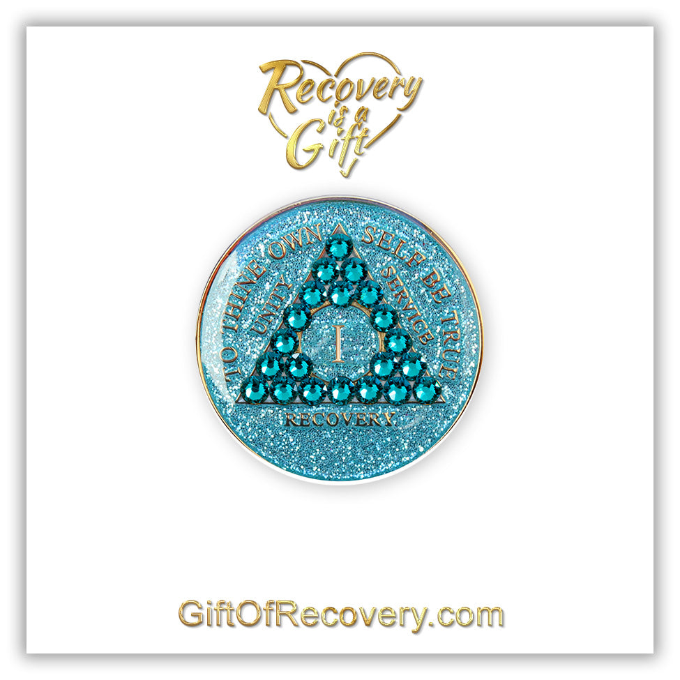 1 year Aqua glitter AA medallion on a white 3x3 card, with recovery is a gift going through a heart on the top and giftofrecovery.com on the bottom in the color gold, the recovery medallion has 21 genuine blue zircon crystals in the shape of the triangle in the center, and to thine own self be true, recovery, unity, service, the roman numeral in the center, embossed in 14k gold plated brass, sealed with resin for a shiny finish.