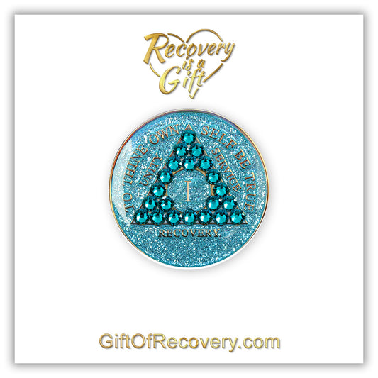 1 year Aqua glitter AA medallion on a white 3x3 card, with recovery is a gift going through a heart on the top and giftofrecovery.com on the bottom in the color gold, the recovery medallion has 21 genuine blue zircon crystals in the shape of the triangle in the center, and to thine own self be true, recovery, unity, service, the roman numeral in the center, embossed in 14k gold plated brass, sealed with resin for a shiny finish. 
