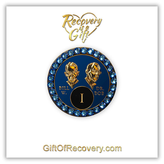 1 year AA Bill & Bob Big Book Blue recovery medallion with 28 genuine Blue crystals around the rim, this medallion is to honor our founders and our time, stylized busts of Bill W. and Dr. Bob, with their names, are embossed 14k gold-plated brass, the medallion is sealed with resin for a glossy finish. seen on a white 3x3 card with recovery is a gift going through an open heart at the top and giftofrecovery.com on the bottom, both in the color gold.