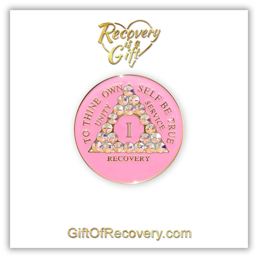 1 Year AA medallion princess pink with 21 Aurora Borealis CZ genuine crystals symbolizing beauty and change from working the program, AA slogan and 3 legacies are embossed with 14k gold-plated brass, sealed with resin for a lasting finish, seen on a white 3x3 card with recovery is a gift going through an open heart at the top and giftofrecovery.com at the bottom, both in the color gold.
