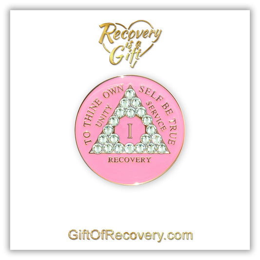 1 year AA medallion princess pink the triangle is adorned with 21 diamond CZ genuine crystals that represent transformation under pressure, the AA slogan and three legacies are embossed with 14k gold-plated brass, the recovery medallion is sealed with resin for a lasting finish, set on a white 3x3 card with recovery is a gift going through an open heart at the top and giftofrecovery.com on the bottom, both in the color gold.