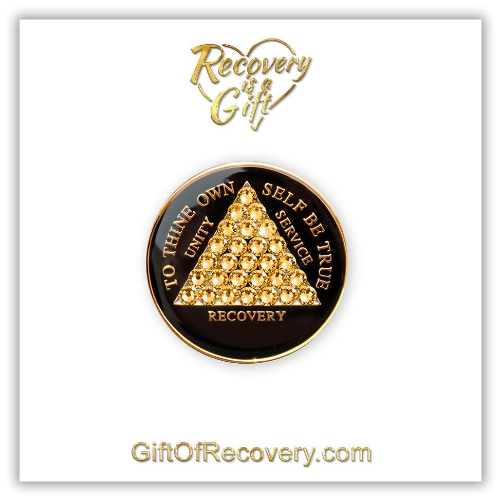 AA medallion black Onyx with 28 gold CZ genuine crystals that form the triangle entirely, this is the timeless recovery medallion, shine on with this beauty, we all have one day at a time, the AA slogan and three legacies are embossed with 14k gold-plated brass and sealed with resin for a glossy finish that will last and is scratch proof, pictured on a white 3x3 card with recovery is a gift going through an open heart at the top and giftofrecovery.com at the bottom, both in the color gold.