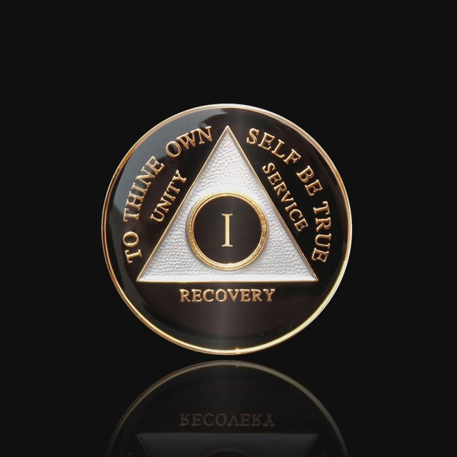 1 year AA medallion Black Onyx, triangle is pearl white, to thine own self be true, unity, service, recovery, and roman numeral are embossed with 14k gold-plated brass, the recovery medallion is sealed with resin for a glossy finish that will last and is scratch free, featured in a 15 second video with a black background.