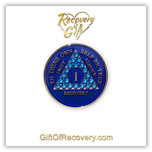 1 year AA medallion Big Book blue adorned with 21 genuine blue CZ crystals symbolizing the importance of our Big Book, to thine own self be true, unity, service, recovery are embossed with 14k gold-plated brass and sealed with resin for a glossy finish that is scratch proof and will last, seen on a white 3x3 card with recovery is a gift going through an open heart at the top and giftofrecovery.com on the bottom, both in the color gold.