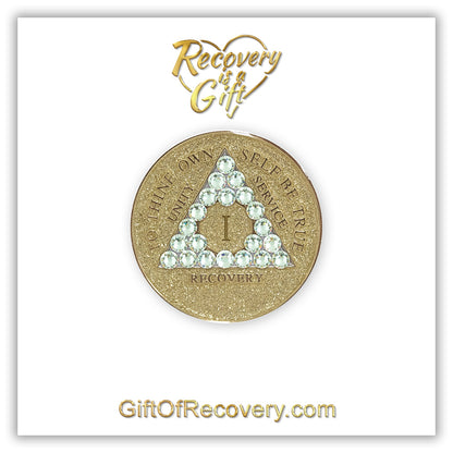 1 year AA medallion in gold glitter with twenty-one genuine clear CZ crystals in the shape of a triangle, symbolizing transformation under pressure, to thine own self be true, unity, service, recovery and roman numeral are embossed with 14k gold-plated brass along with outer rim, sealed in a chip and scratch-resistant resin giving it a beautiful glossy look, recovery medallion is featured on a white 3x3 card with recovery is a gift going through a heart at the top and giftofrecovery.com at the bottom. 