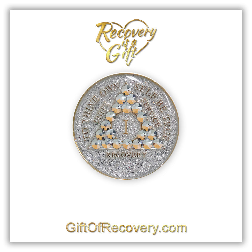 1 year silver glitter AA medallion with 21 genuine comet crystals making up the triangle, the year, unity service, recovery, and to thine own self be true, are embossed in 14k gold along with the rim of the medallion, entirely hand painted and sealed with resin for a glossy finish, set on a white 3x3 card with the lettering in the color gold.