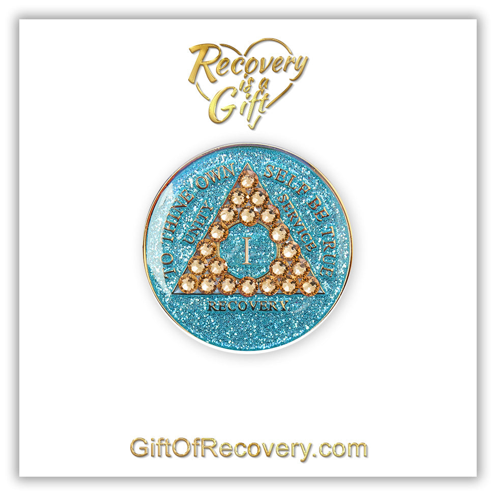 1 year Aqua glitter AA medallion with 21 gold genuine crystal making a triangle in the center, and to thine own self be true, unity, service, recovery, the roman numeral in the center and the rim of the recovery medallion embossed in 14k gold plated brass, the medallion is on a white 3x3 card with recovery is a gift in gold writing going through a gold heart.